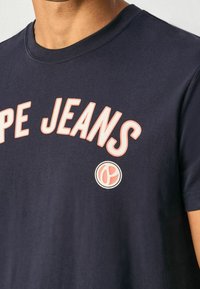 Dunkelblaue Baumwollt-shirt mit dem Aufdruck "Pepe Jeans" in fetter pinker Schrift und einem kreisförmigen Logoakzent. Kurzärmlig und mit Rundhalsausschnitt.