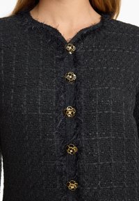 Zwarte gestructureerde jas met een fuzzy rand, voorzien van zes gouden bloemvormige knopen met zwarte centra langs de voorkeursopening.