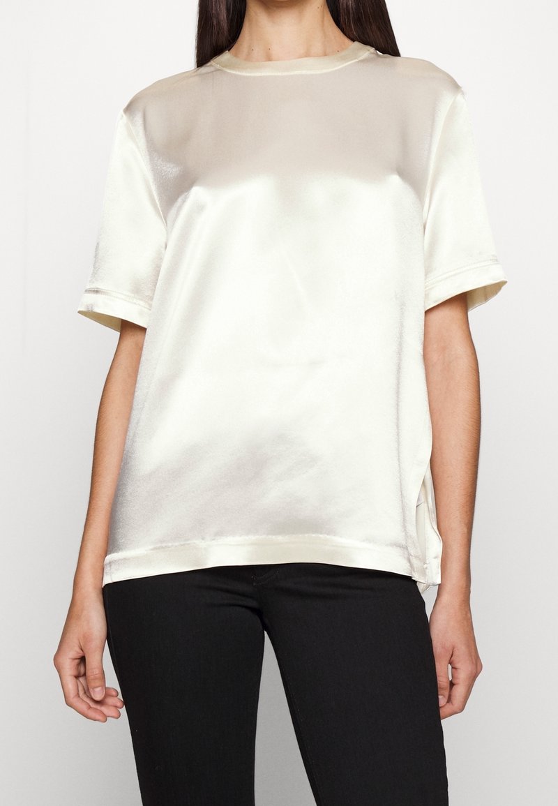 Blusa in satin a maniche corte col colore bianco sporco, con scollo rotondo, fessure laterali e una superficie liscia, abbinata a jeans neri.