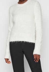 Kvinna iklädd en vit fluffig långärmad tröja och svarta åtsittande byxor, stående mot en enkel ljus bakgrund.