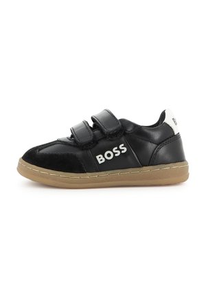 Zwarte leren sneaker met twee klittenbandsluitingen, beige zool en het woord "BOSS" op de zijkant en hielband in witte letters.