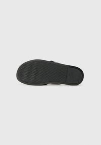 Black sandal bottom view with textured rubber sole; features geometric patterns for grip and a smooth finish around the edges.