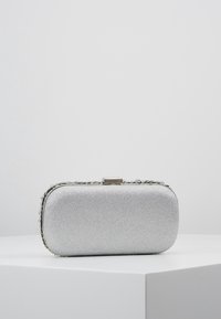 Srebrna kopertówka wykonana z błyszczącego materiału, prostokątna o zaokrąglonych krawędziach, ozdobiona kryształowym wykończeniem i metalowym zapięciem.