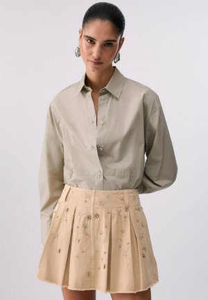 Donna con capelli pettinati all’indietro, che indossa una camicia beige con bottoni e una gonna beige plissettata con decorazioni floreali e perline.