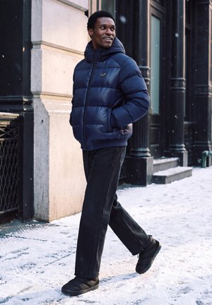 Homme portant une doudoune bleue et un pantalon noir marchant sur un trottoir enneigé dans une zone urbaine, regardant sur le côté et souriant.