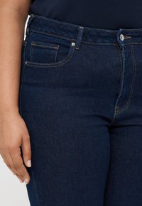 Temno modri kavbojki imajo prilegajoč dizajn, pet žepov in kontrastno šivanje. Pas vsebuje kovinski gumb in zadrgo.