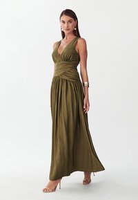 Vrouw draagt mouwloze maxi-jurk in olijfgroen met wikkelbodice en geplooide rok, geaccentueerd met een gouden manchette en pareloorbellen.