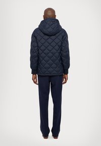 Marineblå quiltet jakke med hætte, med diamantmønstre og elastiske manchetter. Kombineret med marineblå bukser, set fra bagsiden.