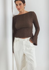 Bruine gebreide lange mouw top met een brede halslijn, gecombineerd met witte hoge taille broeken, gestyled met de handen in de zakken.