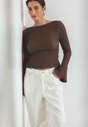 Brown textured long-sleeve top with a wide neckline, paired with white high-waisted pants, styled with hands in pockets.