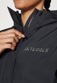 Sort vandtæt jakke med høj krave, lynlåslukning og et broderet TERREX-logo på brystet. Glat, let materiale.