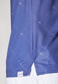 Maglietta sportiva viola con un motivo geometrico, caratterizzata da punti bianchi e dettagli. Include un'etichetta logo sul fondo.