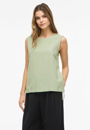 Kobieta w bezrękawniku w jasnym zielonym kolorze i szerokich czarnych spodniach, stoi z jedną ręką w kieszeni na białym tle.