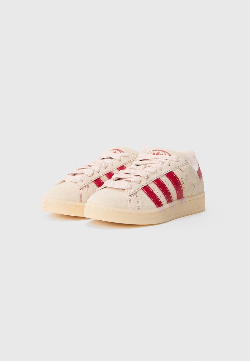 Dos zapatillas deportivas con parte superior de ante beige, detalles rojos, tres rayas rojas en el lateral y suela de goma color tostado. Cordones planos y marca minimalista.