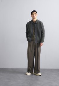 Grå stickad cardigan över en grön skjorta, i kombination med ljusbruna vida byxor och beige skor, stående mot en neutral bakgrund.
