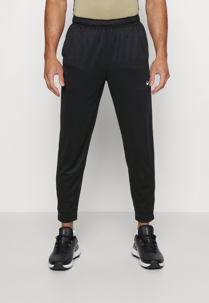 Homme portant un jogging Nike noir et des chaussures de sport noires, debout devant un fond uni.