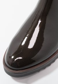 Bout de botte brun foncé brillant avec un motif texturé fin et une épaisse semelle en caoutchouc noire sur fond blanc.