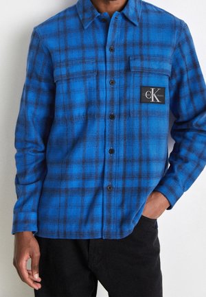 Man wearing a blue and black plaid button-up shirt with a Calvin Klein Jeans logo patch on the chest pocket, paired with black pants.