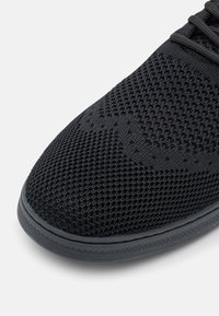 Gros plan sur l’avant d'une basket en tissu tricoté noir avec un motif texturé et une semelle en caoutchouc noir sur fond blanc.