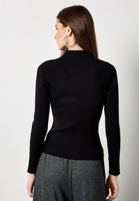 Top lungo a maniche nere a coste con un design aderente e colletto alto. La texture è liscia, abbinata a pantaloni grigi a quadri.