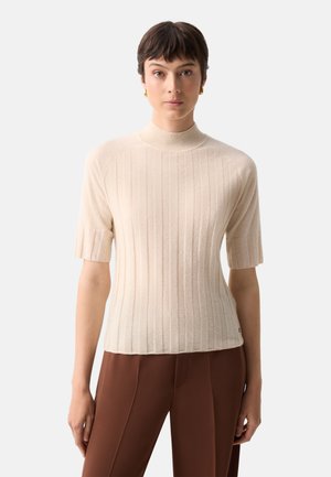 Kvinde med kort mørkt hår, iført en beige ribstrikket kortærmet sweater og brune bukser, stående mod en ensfarvet lys baggrund.