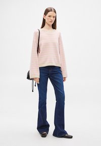 Μαλακό ριγέ ροζ και μπεζ πλεκτό πουλόβερ με στρογγυλή λαιμόκοψη, συνδυασμένο με σκούρα μπλε καμπάνες τζιν. Το μοντέλο κρατά μια μικρή μαύρη τσάντα με κορδόνι.