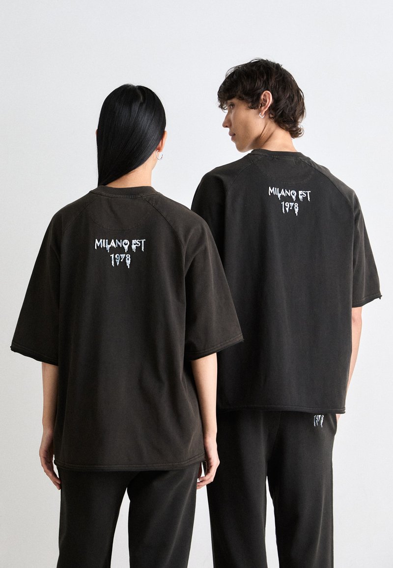 To personer iført sorte oversize t-shirts med hvid "MILANO EST 1978" tekst på den øverste del af ryggen, står foran en ensfarvet hvid baggrund.