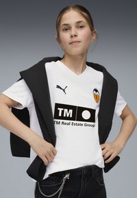 Maglietta bianca a V con maniche corte e logo Puma, constemmazione di Valencia ed un testo sponsorizzato in nero. Felpa nera appesa sulle spalle.