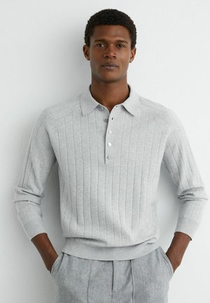 Noor mees, kelle käed on taskutes, kannab helehalli soonikkoelise polo-kampsunit ja sobivaid halli värvi pükse, taustaks valge sein.