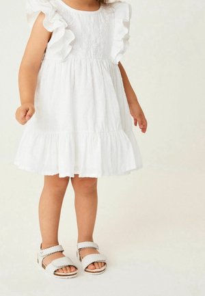Biała sukienka z falbanianymi rękawami, fakturowanym gorsetem i warstwowym dołem, zestawiona z białymi sandałami. Miękki, lekki materiał.