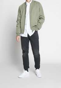 Giacca bomber verde sopra una camicia bianca a bottoni, abbinata a jeans neri strappati e sneakers bianche. Design semplice e vestibilità casual.