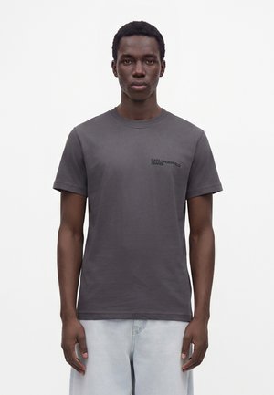 Ung mand med mørk hud iført en ensfarvet mørkegrå T-shirt med teksten "Karl Lagerfeld Jeans" og lyse jeans mod en hvid baggrund.