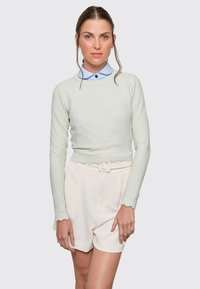 Light green textured sweater with long sleeves, scalloped hem, worn over a light blue collared shirt, paired with white high-waisted shorts.