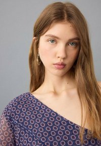 Záběr zblízka osoby s dlouhými hnědými vlasy, která má na sobě modrý vzorovaný top s květinovými motivy a jemné náušnice, na šedém pozadí.