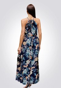 Maxi-jurk in marineblauw met bloemenprint. Kenmerkt zich door een halterneck met strikdetail, vloeiende stof en een losse pasvorm, wat beweging en comfort bevordert.