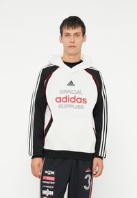 Moletom branco e preto com detalhes em vermelho, apresentando um logotipo oversized da "adidas" e o texto "FORNECEDOR OFICIAL". Três riscas nas mangas.
