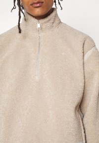 Beige fleece-tröja med hög krage. Har en dragkedja fram till hälften och en subtil logodetalj. Mjuk textur och avslappnad passform.