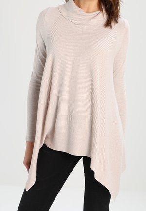 Femme portant un pull-tunique asymétrique à manches longues beige clair avec une texture côtelée et un pantalon noir sur fond uni.