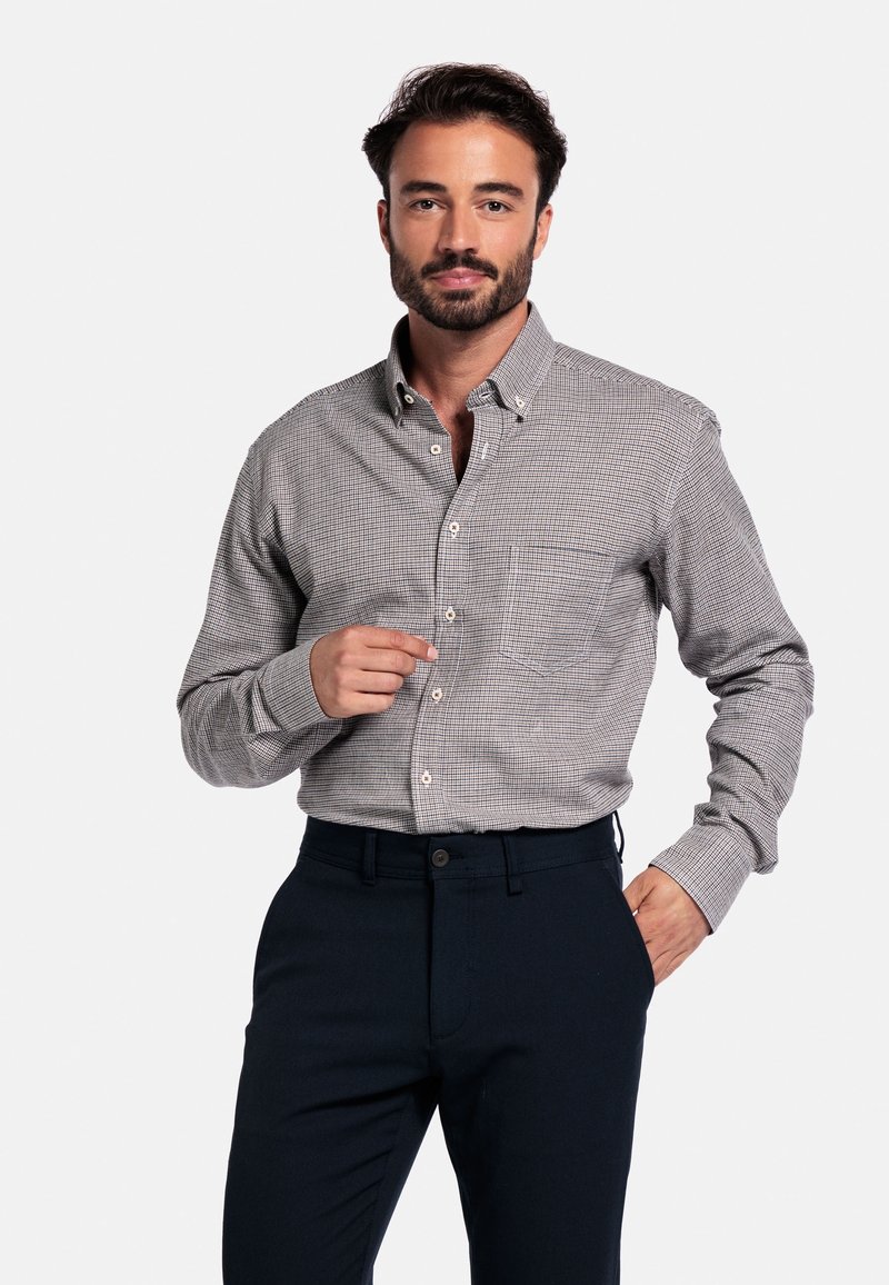 Man met baard in een grijs geruit overhemd en donkerblauwe broek, die zijn overhemd aanpast terwijl hij met één hand in zijn zak zit.