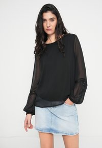 Μαύρο πουκάμισο με διαφανή μακριά μανίκια και λεπτομέρειες δαντέλας. Συνδυάζεται με μίνι φούστα από ανοιχτό μπλε denim. Το μοντέλο στέκεται με τα χέρια στις τσέπες.