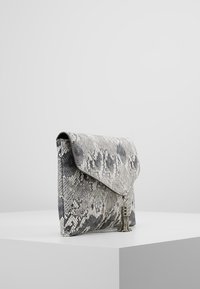 Harmaa-musta käärmekuvioinen clutch-laukku, jossa on läppäkiinnitys, teksturoitu pinta ja koristeellinen metallinen hapsu.