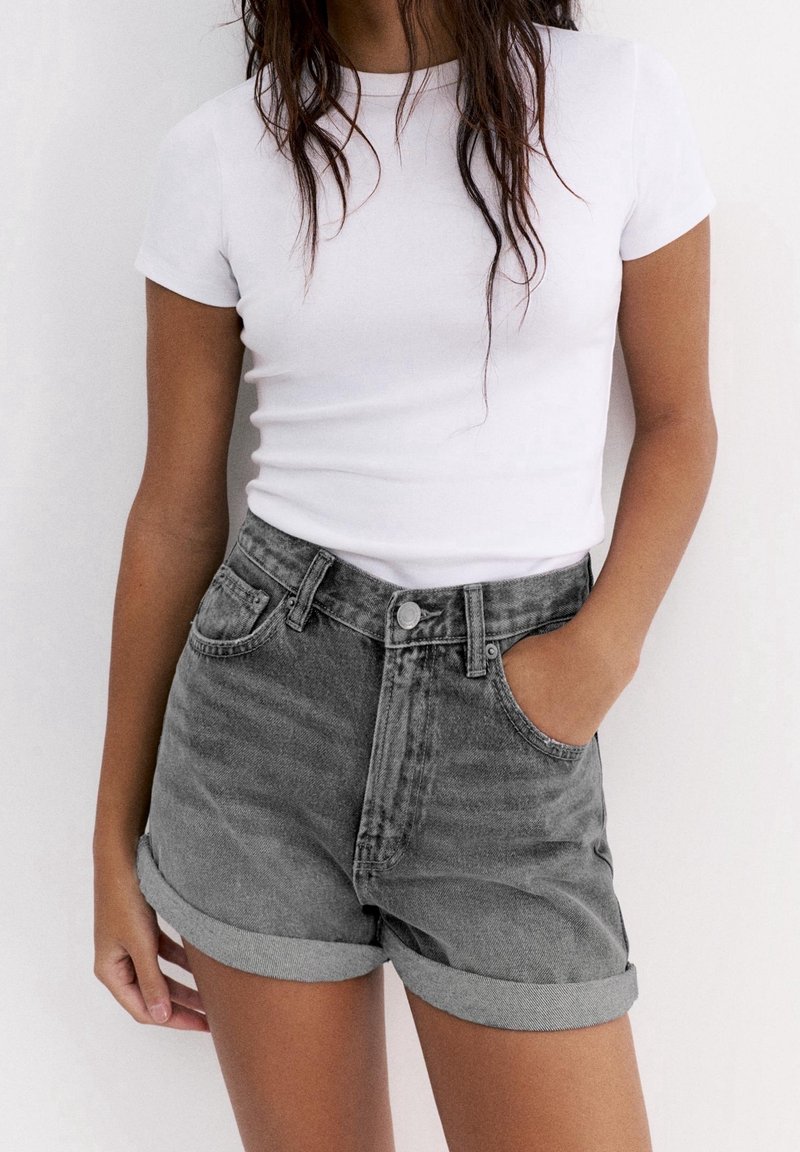 Femme portant un t-shirt blanc simple ajusté, rentré dans un short en jean noir taille haute retroussé, main dans la poche.