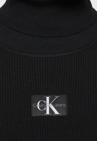 Zwarte geribbelde turtleneck trui met een Calvin Klein Jeans label in het midden. Heeft een hoge halslijn en een gestructureerde stof.