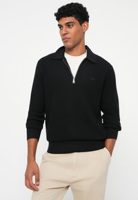 Schwarzer gerippter Pullover mit Reißverschlusskragen, langen Ärmeln und taillierter Passform. Mit einem kleinen schwarzen Logo auf der Brust. Über einem weißen Hemd getragen.
