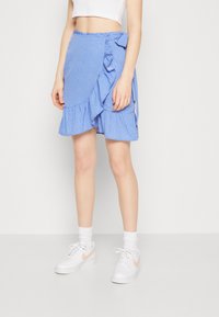 Jupe bleu à pois blancs, avec des détails en volants et un nœud sur le côté. Portée avec des baskets blanches et des chaussettes de cheville.