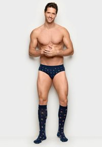 Muž bez košile, oblečený v tmavě modrých slipách a podkolenkách s motivy perníčků a cukrových hůlek, usmívající se se sepnutýma rukama.