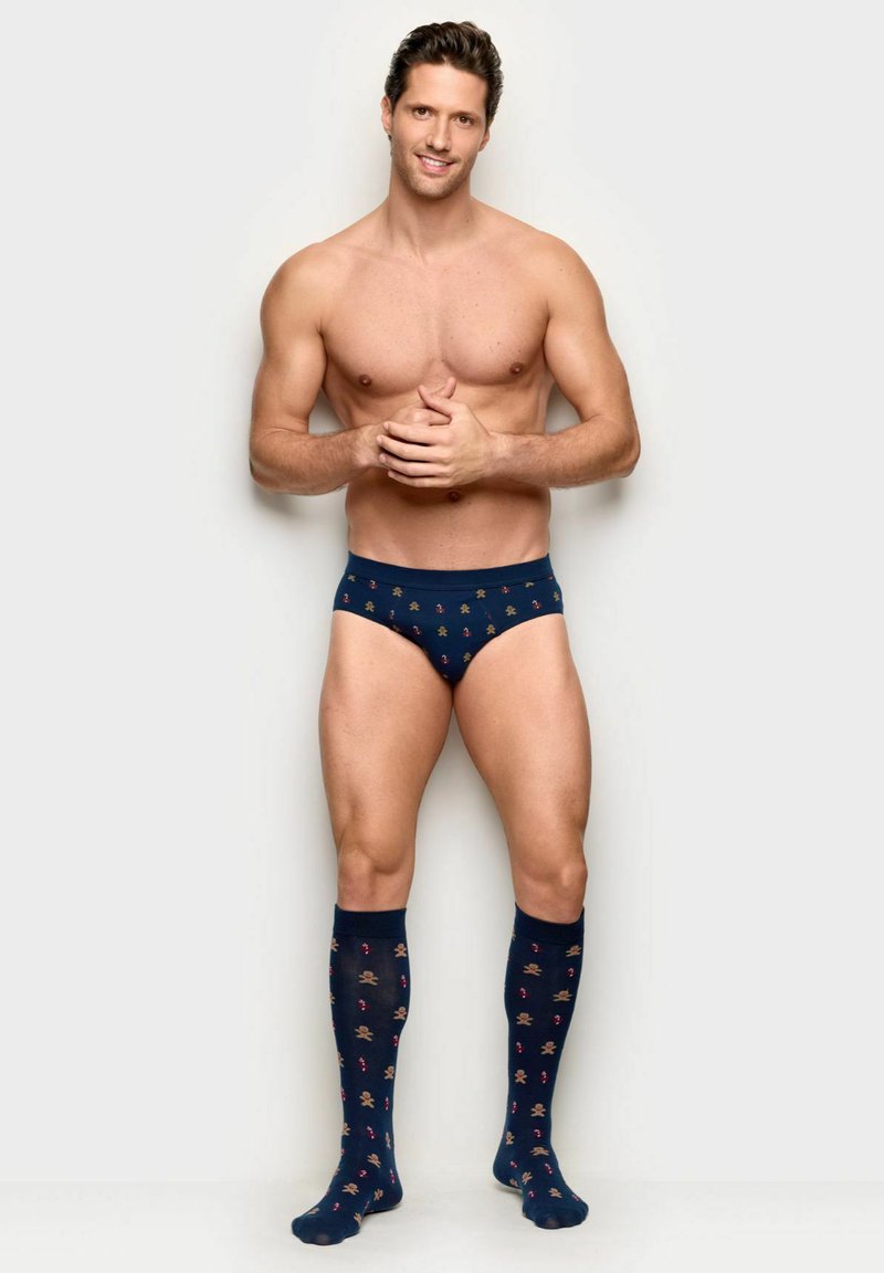 Muž bez košile, oblečený v tmavě modrých slipách a podkolenkách s motivy perníčků a cukrových hůlek, usmívající se se sepnutýma rukama.