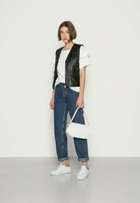 Melnas ādas veste, balts viļņots T-krekls, zili džinsi, balti sporta apavi, maza balta rokassoma; modele stāv pret neitrālu fonu.