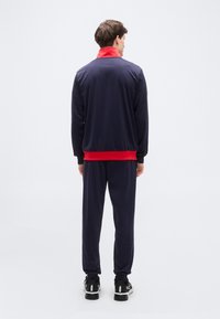 Φόρμα με μπουφάν σε σκούρο μπλε χρώμα και κόκκινο γιακά, ταιριαστό παντελόνι σε σκούρο μπλε και μαύρα αθλητικά παπούτσια. Το ύφασμα φαίνεται λείο και ελαφρύ.