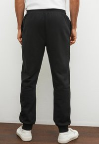 Sort sweatpants lavet af blødt stof med elastisk talje, sidelommer og tapered ben, parret med hvide sneakers på et trægulv.